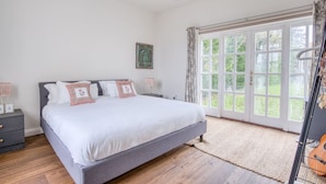 A bedroom with a large bed, a nightstand, a lamp, a guitar, and a view of greenery through the window.