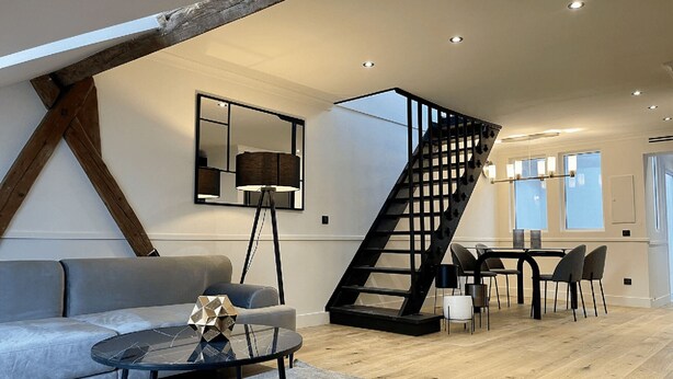 A modern living room with a sofa, coffee table, and a geometric decorative piece. A black staircase with a wooden frame leads to a dining area with a table and chairs. - Alfred Suites is an elegant, upscale residence ideally located in the vibrant he (Dijon)