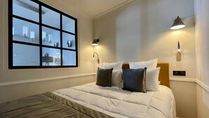A neatly made bed with a grey and white duvet, two bedside lamps, and a large window with a view of the room's interior. - Alfred Suites is an elegant, upscale residence ideally located in the vibrant he (Dijon)