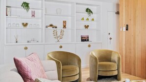 Una sala moderna con dos sillones dorados, un sofá blanco con un cojín rosa y una estantería blanca con adornos y libros.