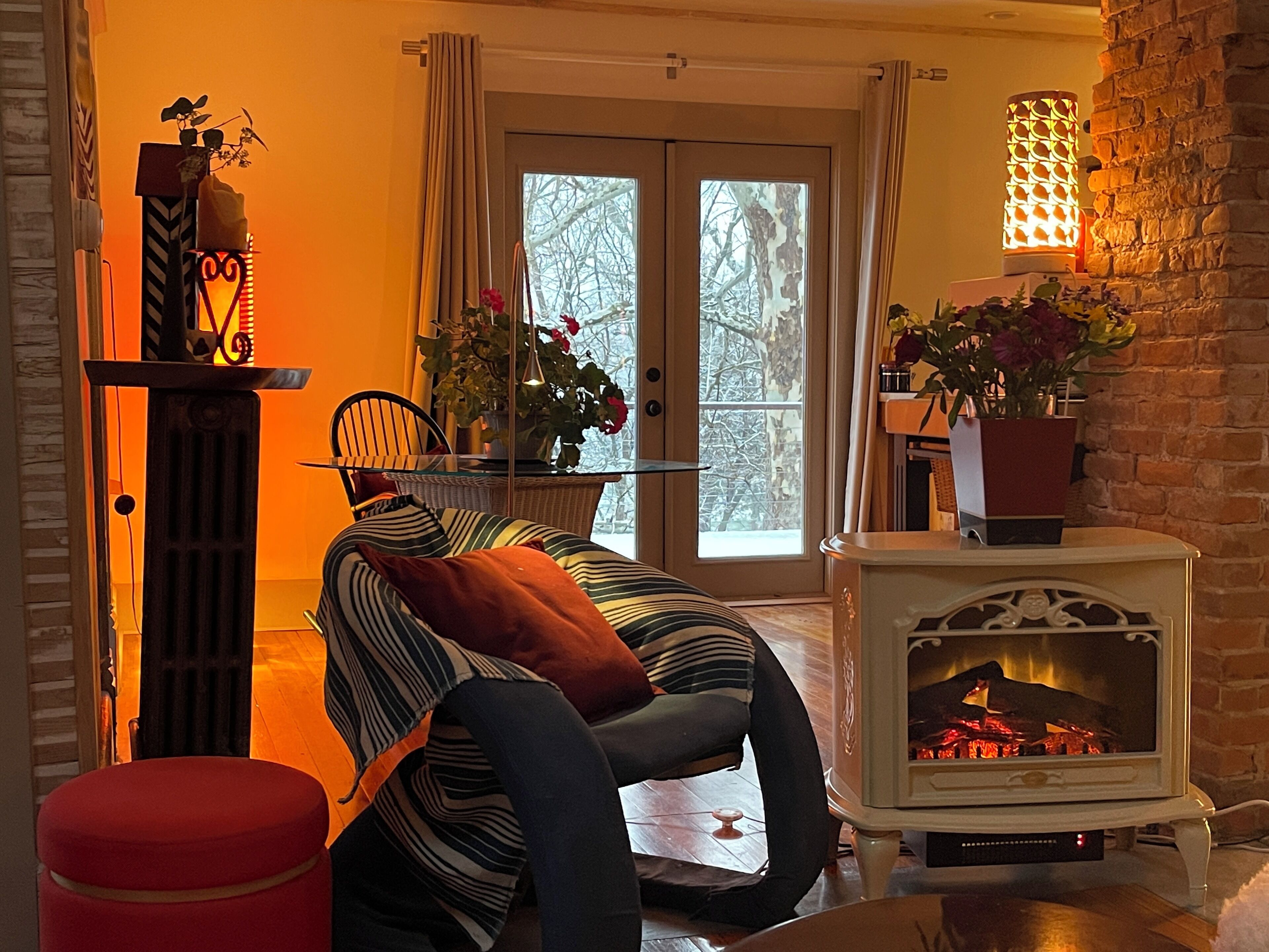Ein gemütliches Wohnzimmer mit Kamin, einem gestreiften Sofa, einem roten Hocker und einer Essecke mit Tisch und Stühlen.