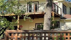 Una casa de dos pisos con un balcón, una terraza de madera y un árbol en primer plano.