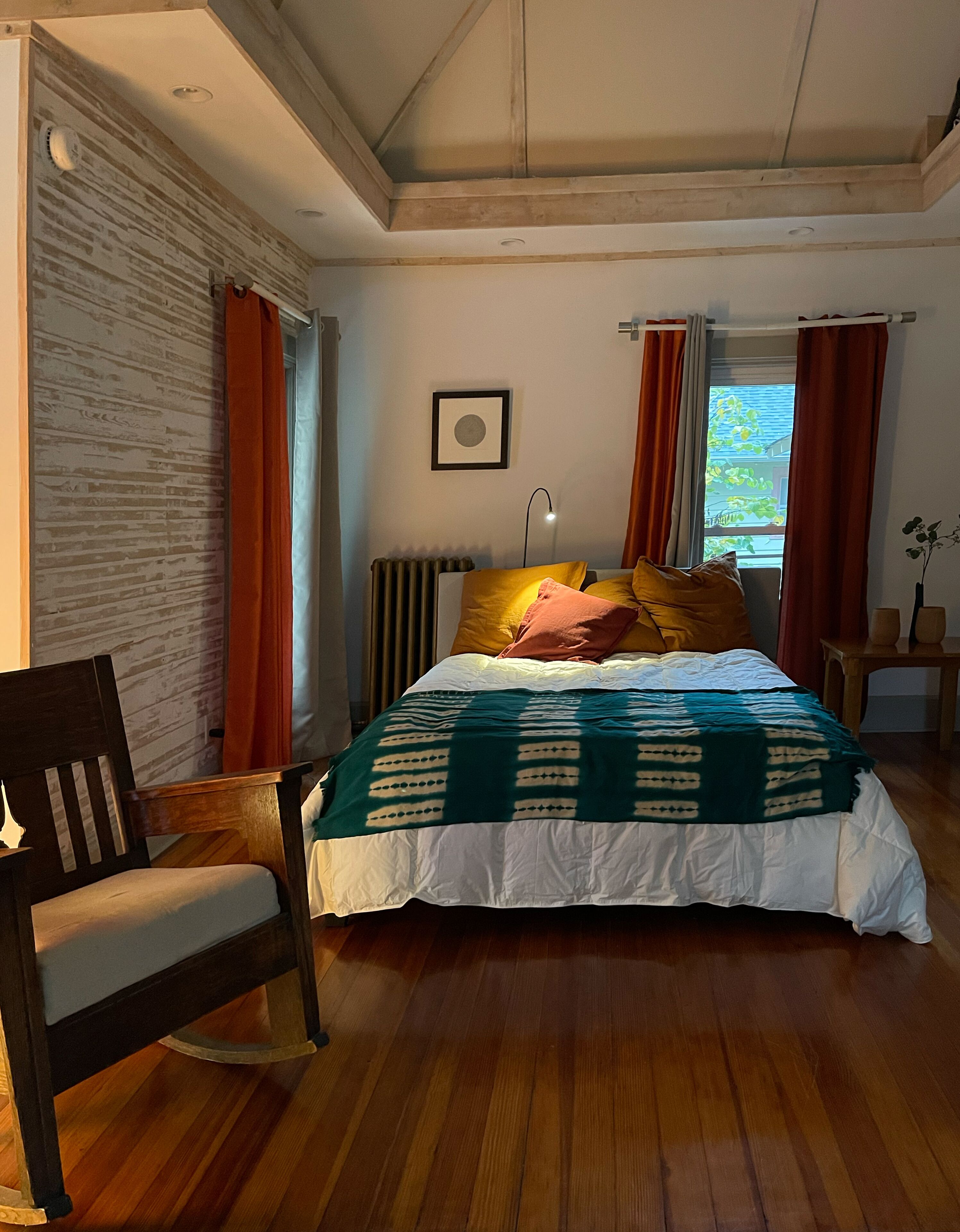 Ein Schlafzimmer mit Holzboden, einem Bett mit gemustertem Bettbezug, einem Sessel, einem Nachttisch, einem Heizkörper und einem Fenster mit Vorhängen.