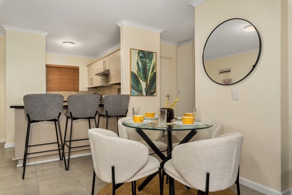 Un coin repas avec une table ronde en verre, quatre chaises grises et un miroir au mur.