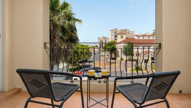 A balcony with two wicker chairs, a small table with a tray of food and drinks, and a view of a street with buildings and palm trees. - Villa Italia Getaway,  Pool, Gym, Netflix, Wifi (Cape Town)