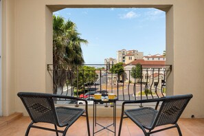 Un balcon avec deux fauteuils en osier, une petite table sur laquelle se trouve un plateau avec de la nourriture et des boissons, et une vue sur une rue bordée d’immeubles et de palmiers.