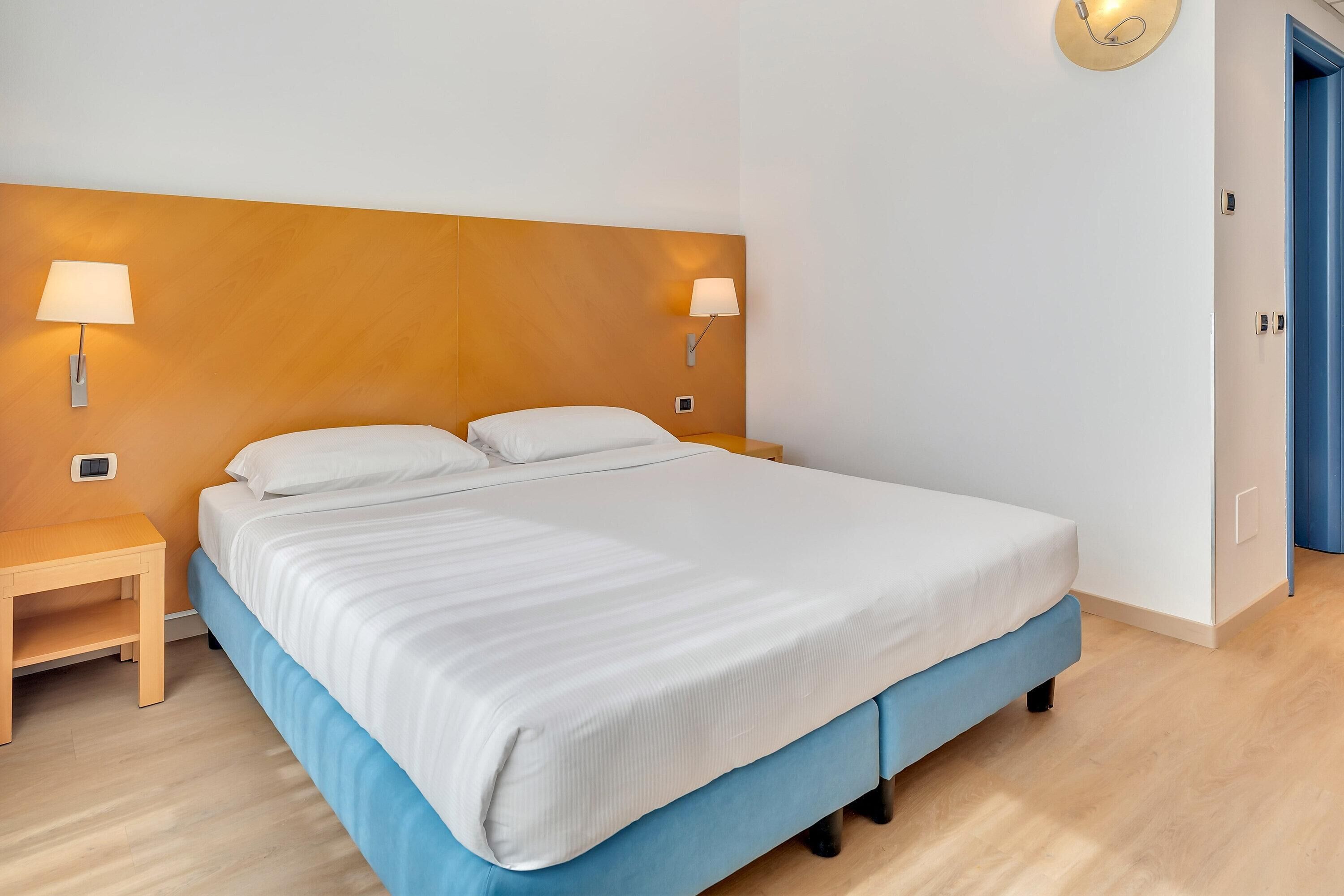 Una habitación de hotel moderna con una cama grande, dos lámparas de noche, una mesita de noche de madera y una puerta azul.
