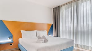 Habitación de hotel con cama, mesita de noche, termostato en la pared y ventana con cortinas.