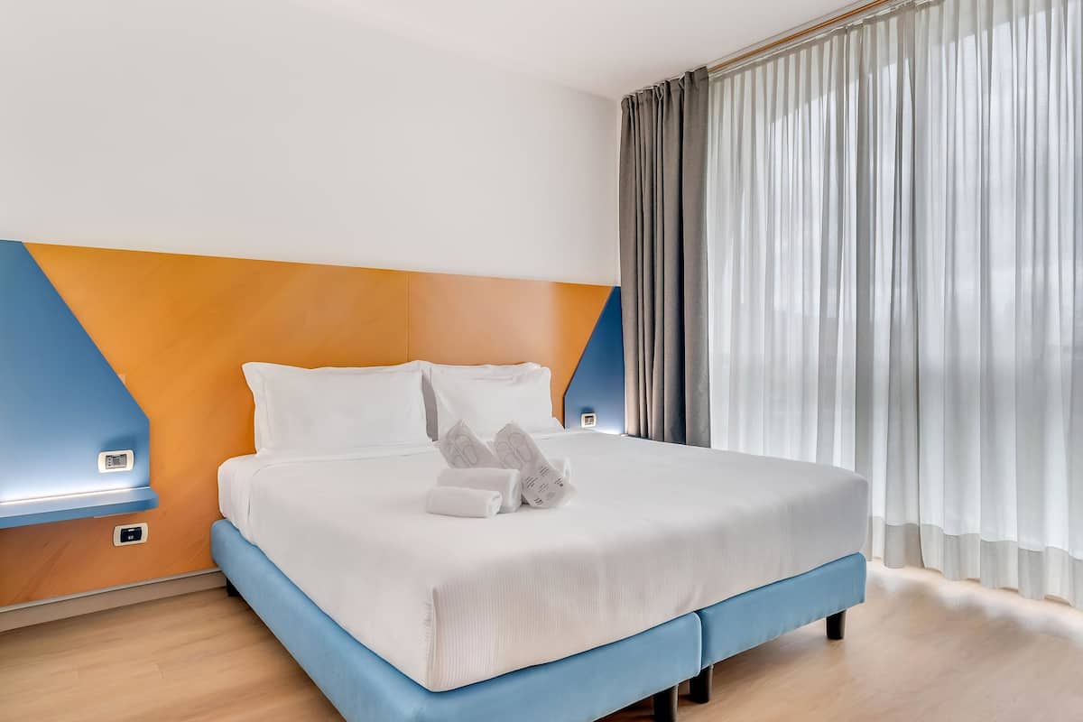 Habitación de hotel con cama, mesita de noche, termostato en la pared y ventana con cortinas.