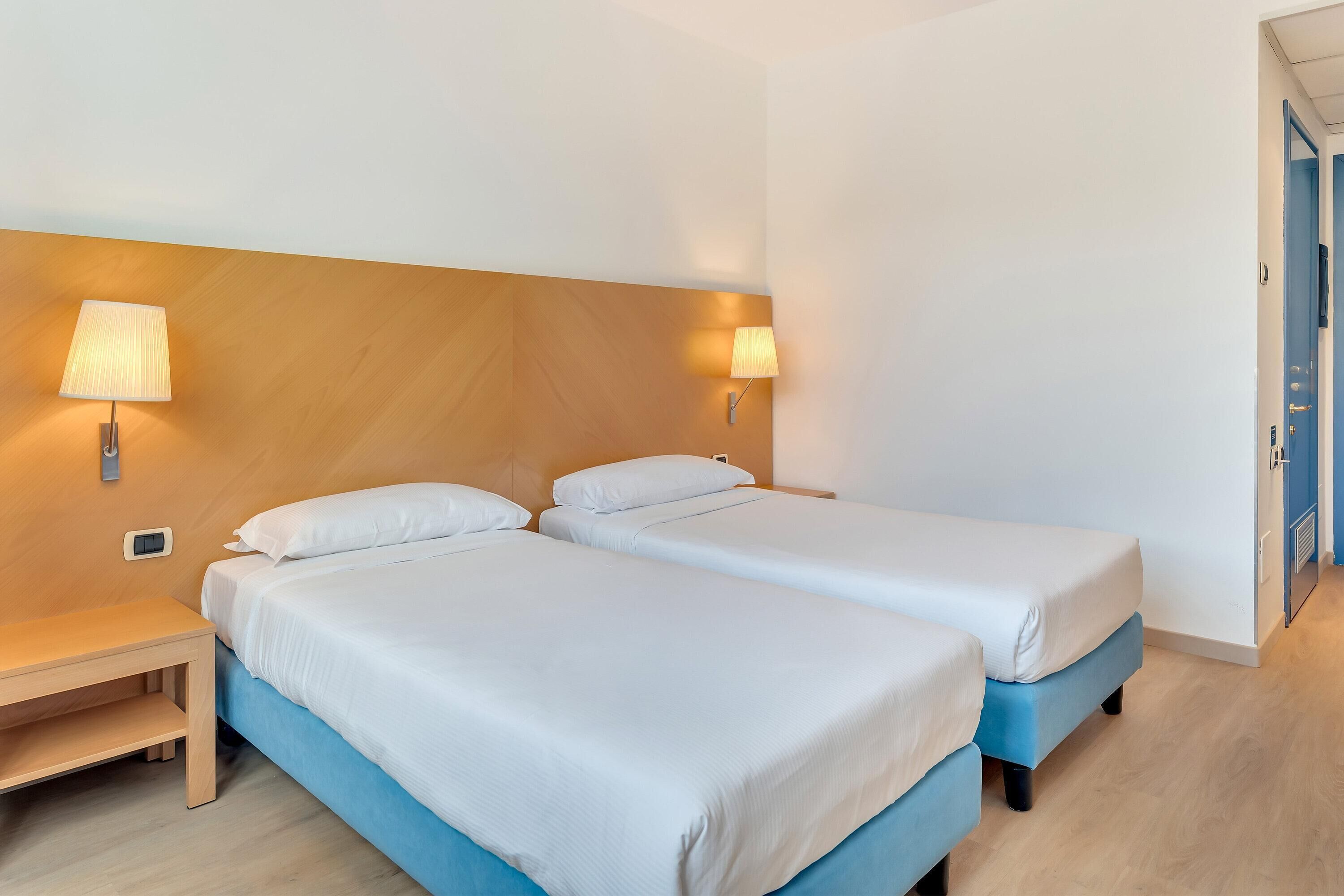 Una habitación de hotel con dos camas, cabecero de madera, lámparas de noche y una mesita de noche pequeña de madera.