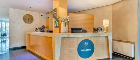 Un mostrador de recepción de hotel con un cartel que dice 'WELCOME', una pantalla digital y un jarrón con una planta.