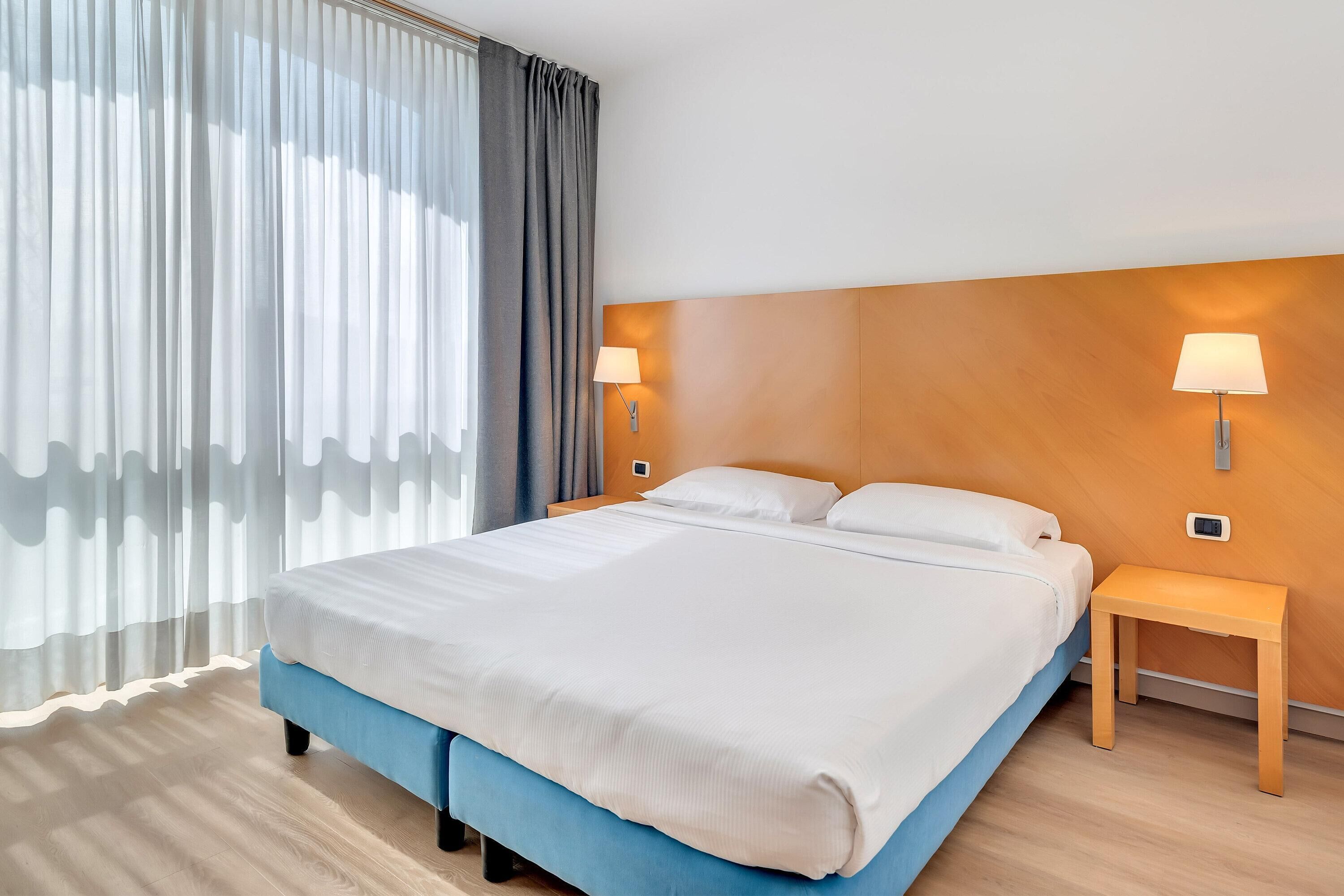 A hotel room with a large bed, two bedside lamps, a wooden headboard, and a small wooden nightstand.