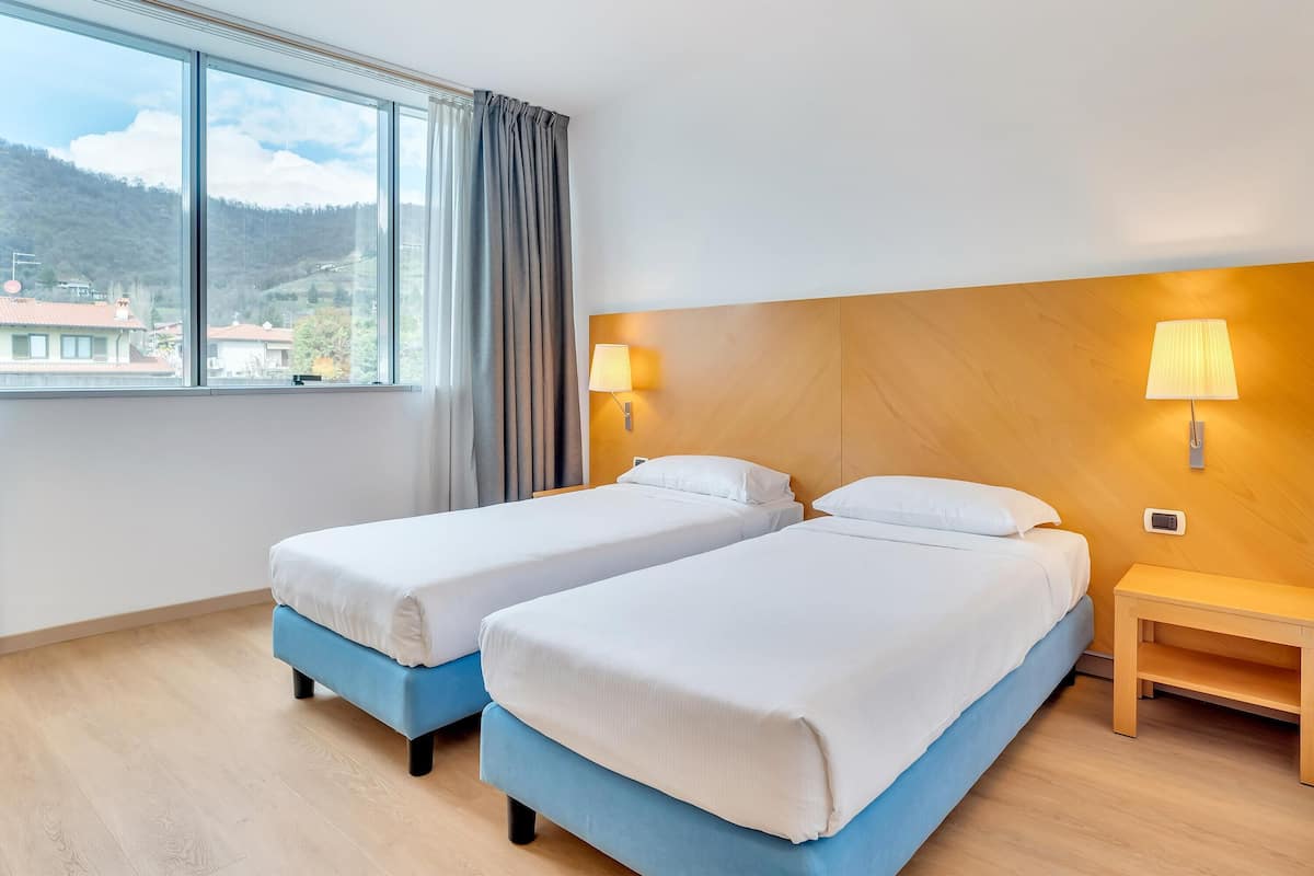 Habitación de hotel con dos camas, un cabecero de madera, una mesita de noche y una ventana con vistas a una montaña y edificios.