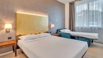 Habitació d'hotel amb un llit gran, un altre més petit, dues taules de nit i una finestra amb cortines.