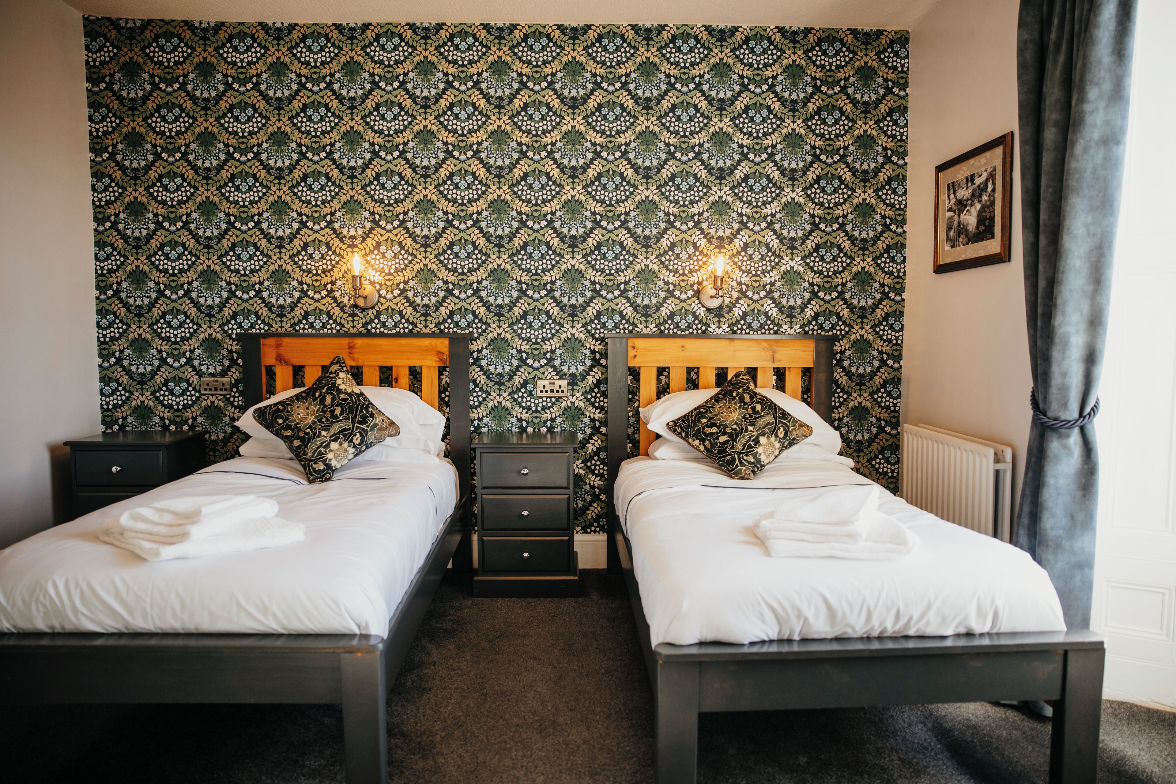 A hotel room with two beds, each with a wooden headboard and white bedding. There are two bedside tables with lamps, and a patterned wallpaper on the wall.