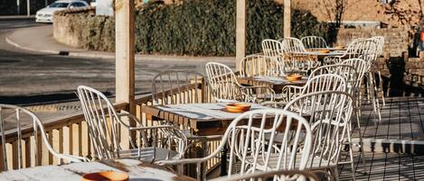 Des tables et des chaises sont disposées à l'extérieur, un bâtiment en briques se dresse à l'arrière-plan et le ciel est dégagé.