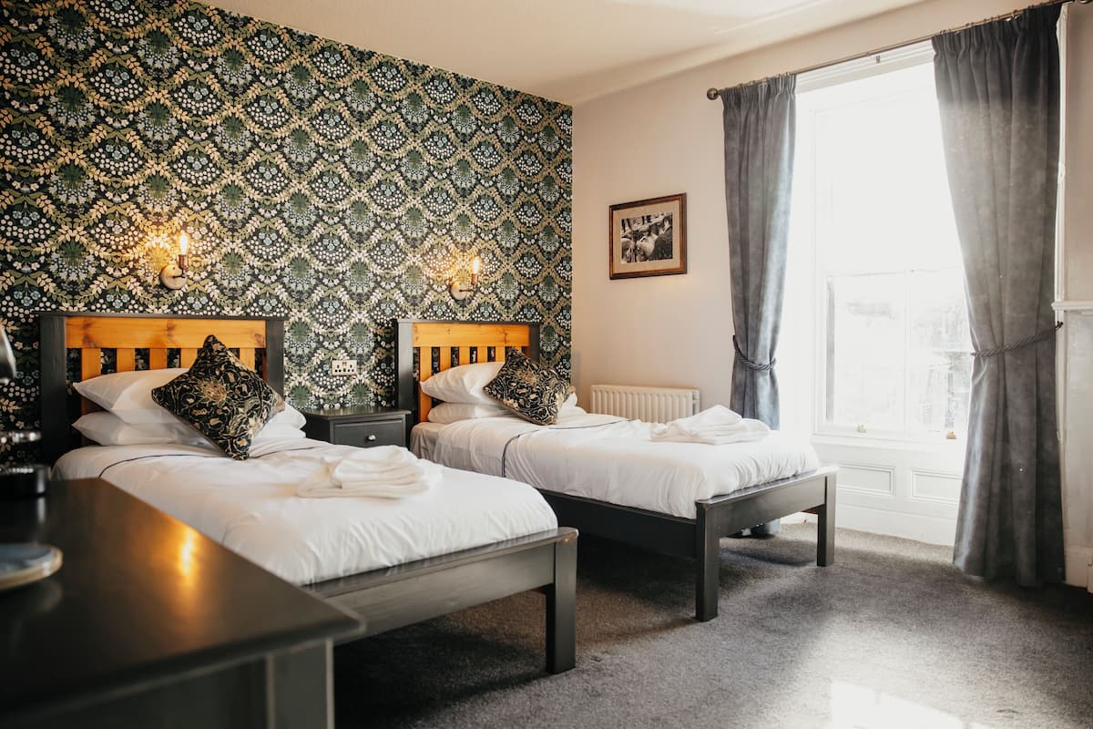 A hotel room with two beds, a nightstand, a table, a radiator, and a window with curtains.