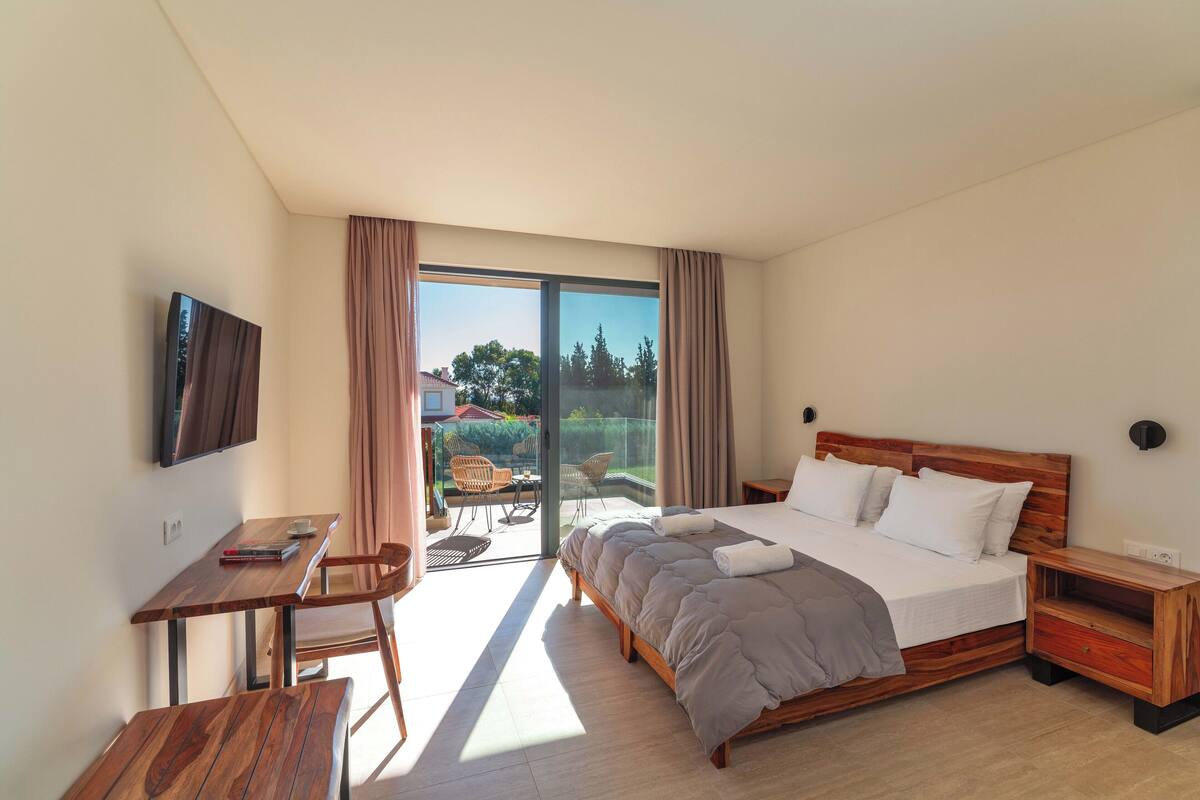 Une chambre d’hôtel moderne dotée d’un grand lit, d’un bureau et d’un balcon offrant une vue sur les arbres et une maison.
