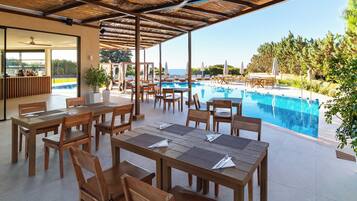A poolside area with wooden tables and chairs, a view of the pool, and a covered seating area with umbrellas.