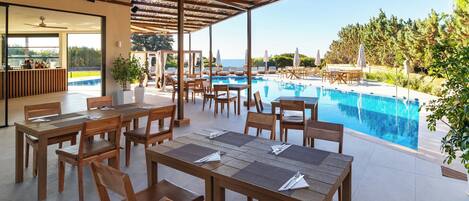 Un espace aménagé au bord de la piscine, avec des tables et des chaises en bois, une vue sur la piscine et un coin salon couvert équipé de parasols.