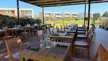 Un espace de restauration en plein air avec des tables et des chaises en bois, un toit de chaume et une vue sur des bâtiments modernes en arrière-plan.