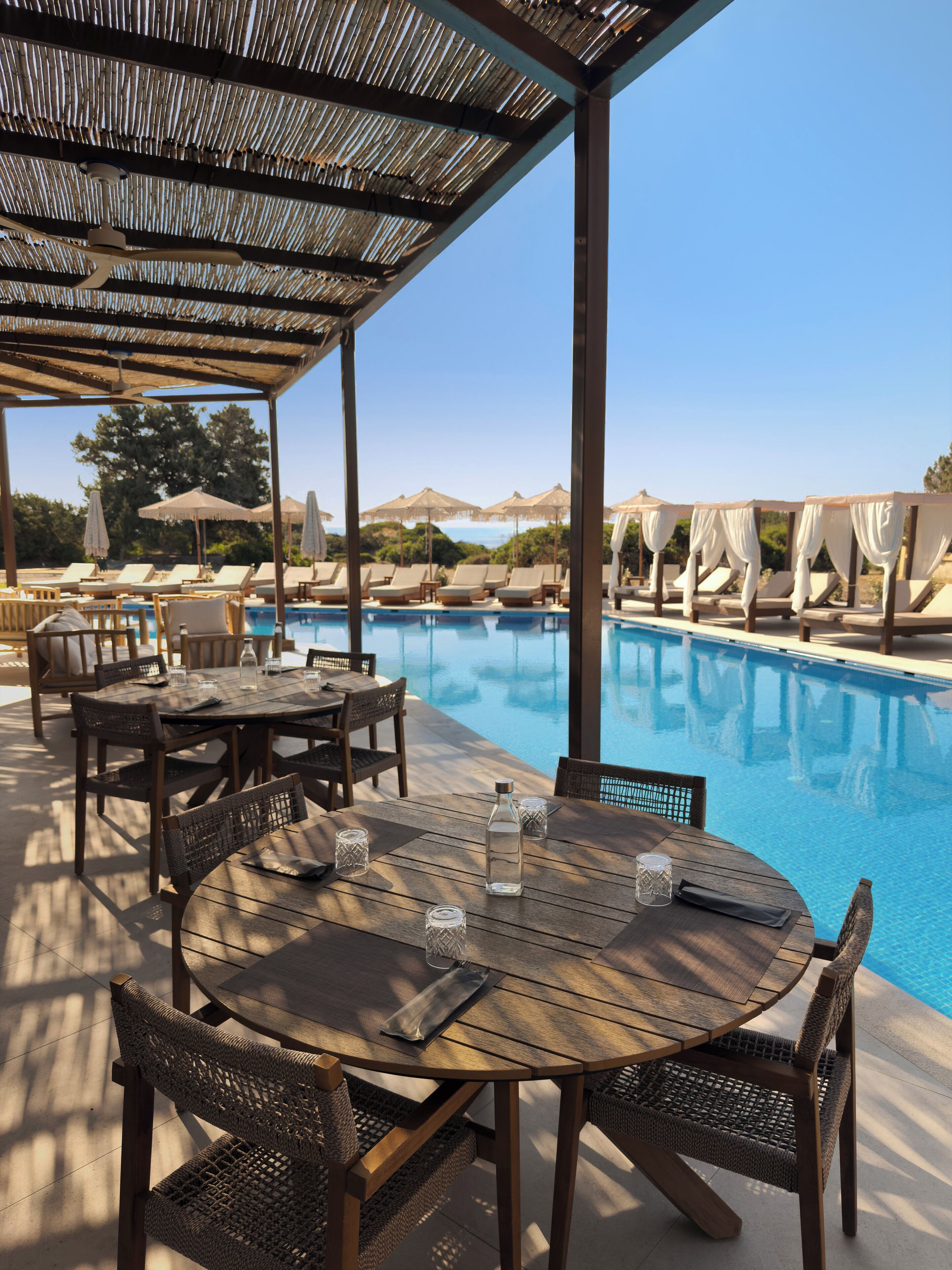 A poolside area with a wooden table and chairs, umbrellas, and lounge chairs.