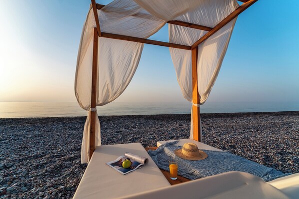 Eine Strandlounge mit einer Holzpergola, einem Tisch mit einer Zeitschrift, einem Hut und einem Glas Orangensaft.