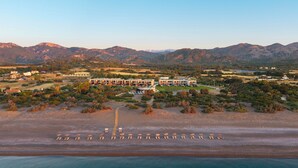 An aerial view of a resort with a beachfront, a large building with multiple floors, and surrounding hills. - Aethrion Luxury Villas & Suites (Rhodes)