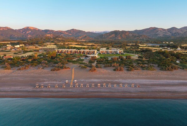 Luchtbeeld van een resort met een strand, een groot gebouw met meerdere verdiepingen en omliggende heuvels.