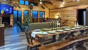 Ein gemütliches Blockhaus-Interieur mit einem Holztisch, der für eine Mahlzeit gedeckt ist, einer Weihnachtstanne und einem Fernseher, der verschiedene Sender zeigt.