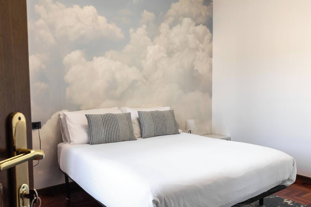 A hotel room with a bed, two pillows, a nightstand, and a wall mural of clouds.