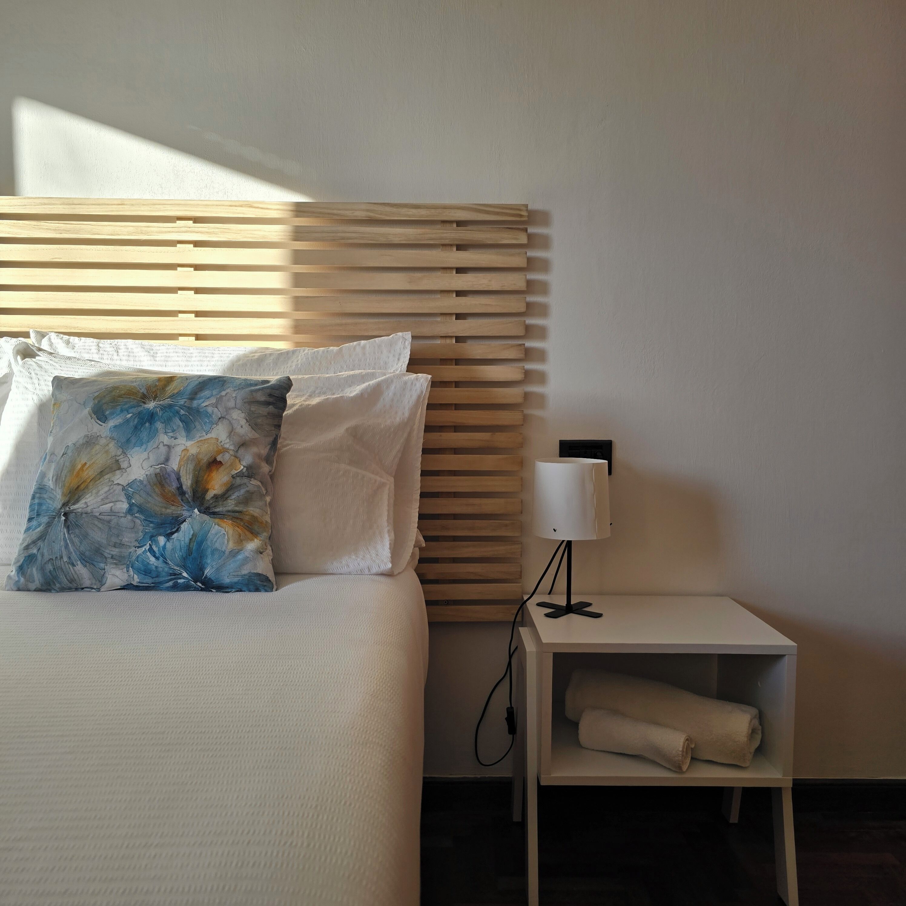 A neatly made bed with a floral pillow, a bedside lamp, and a shelf with a rolled towel.