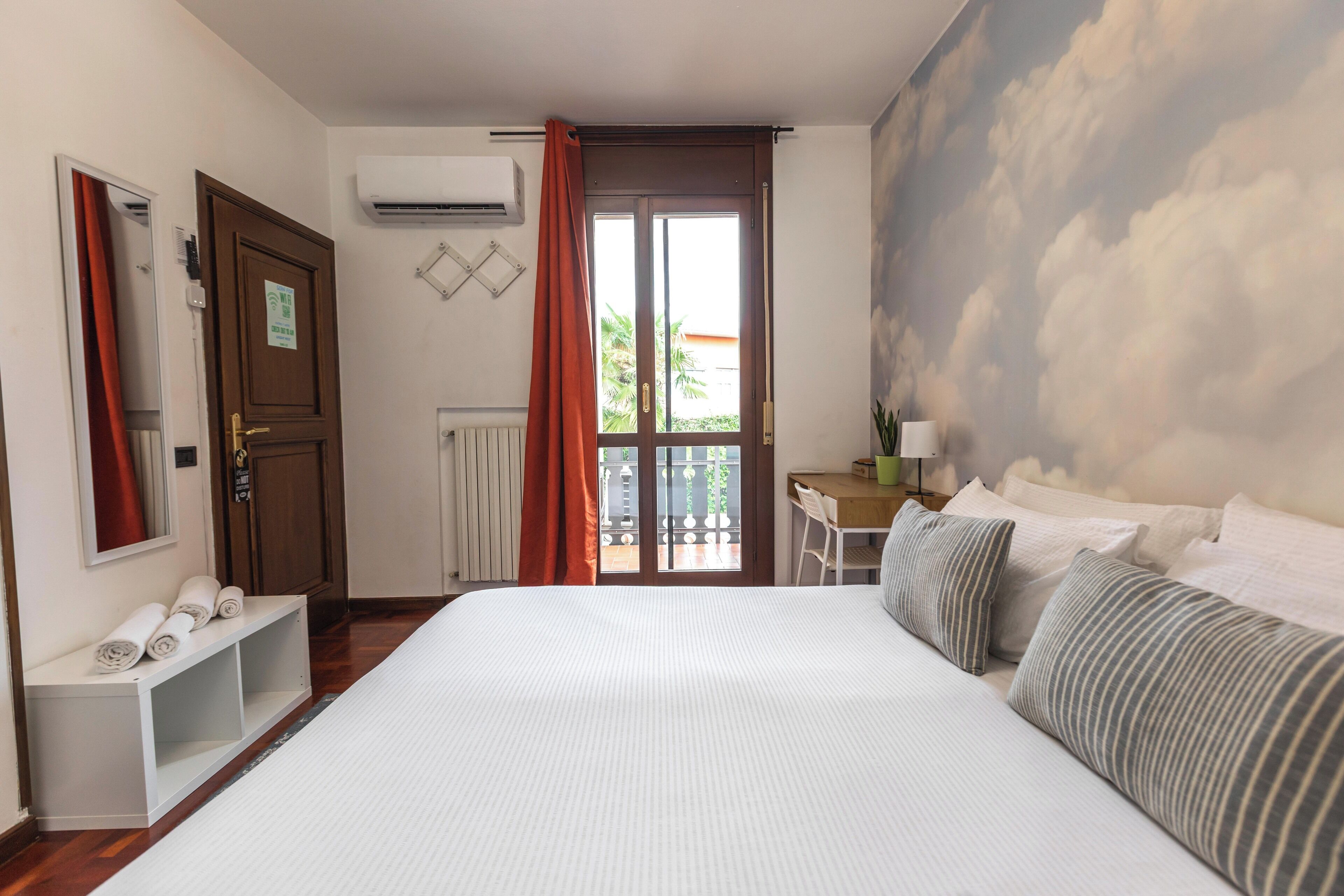 A hotel room with a large bed, a desk, a chair, a window with curtains, a radiator, and a wall-mounted air conditioner.