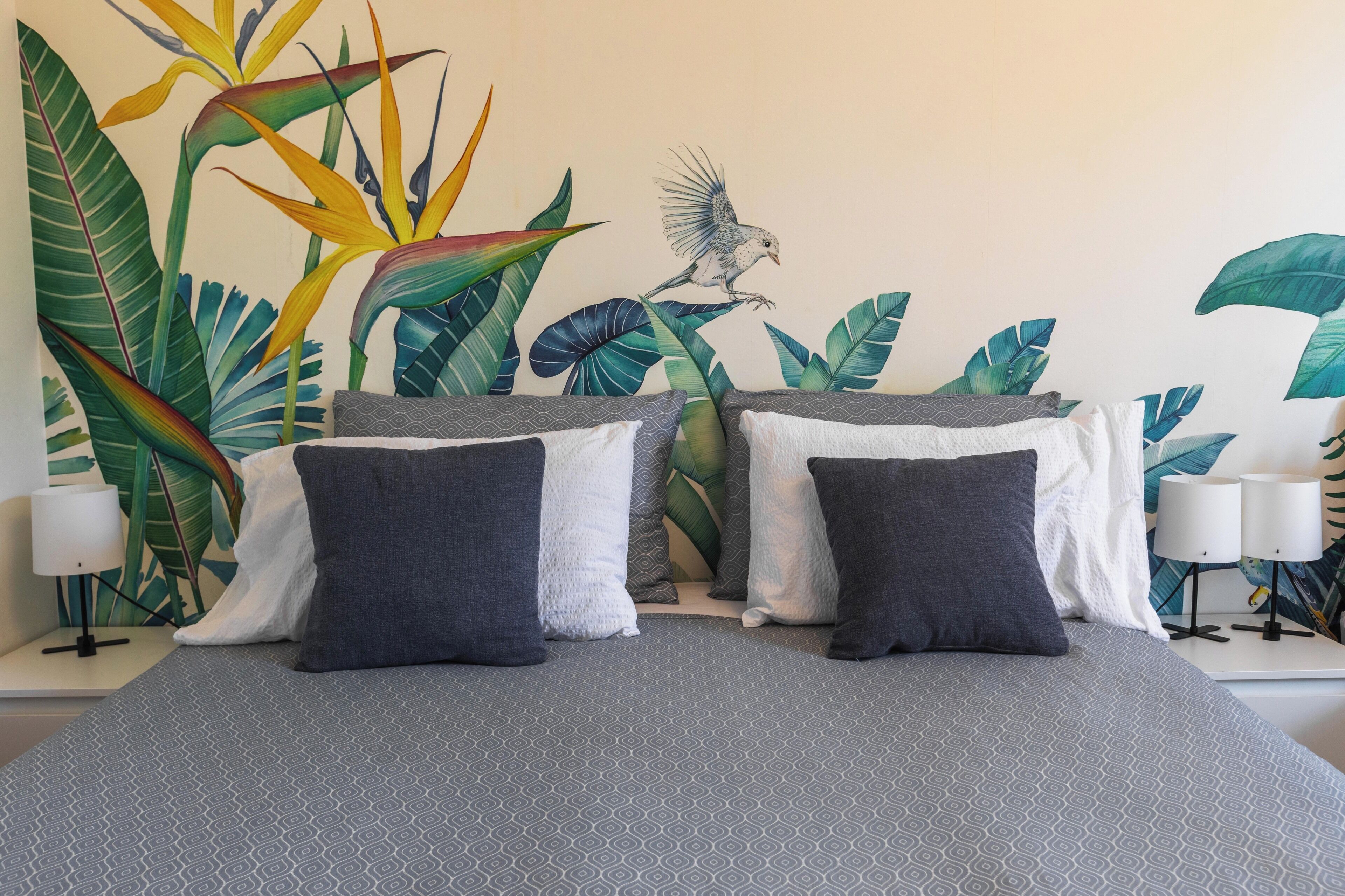 A bed with a patterned gray bedspread, two pillows, and a wall mural of tropical plants and a bird.