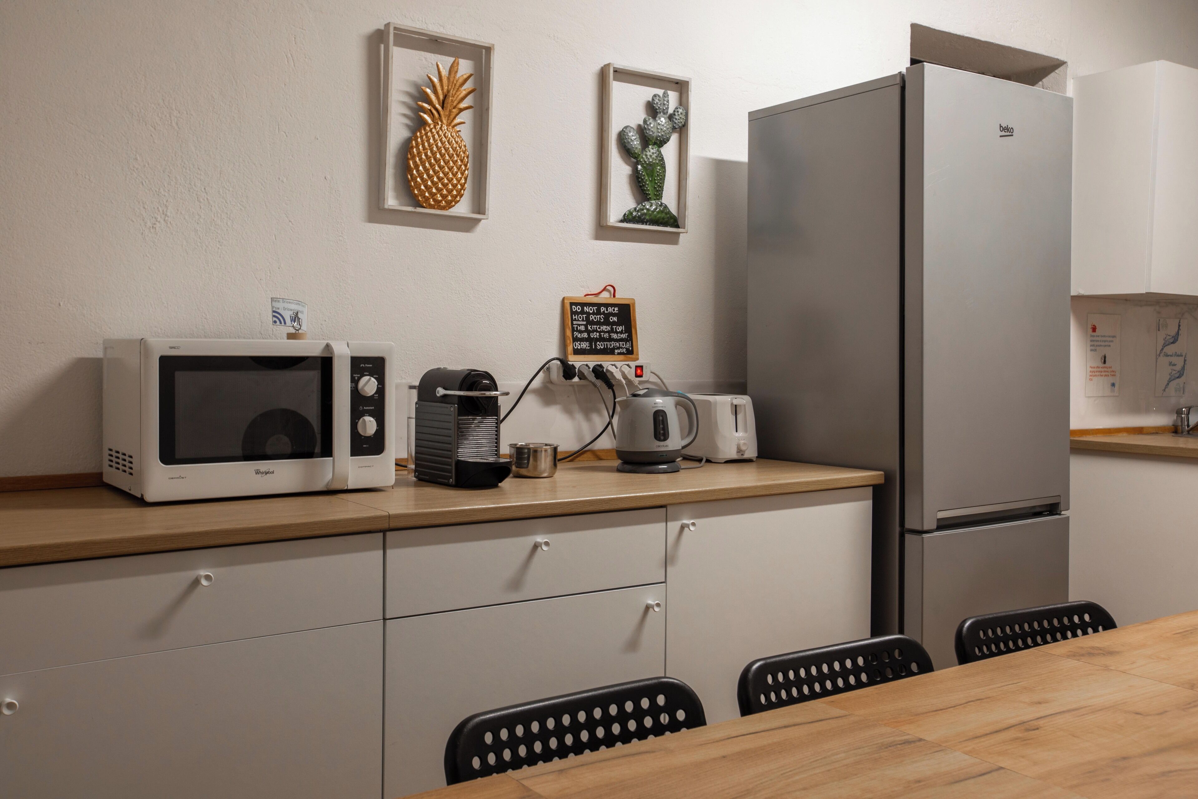 Eine moderne Küche mit Mikrowelle, Kaffeemaschine, Toaster und Kühlschrank. Um einen Holztisch stehen Stühle, an der Wand befinden sich Dekorationen.