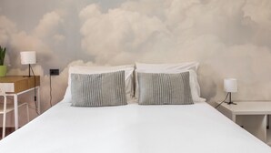 Ein Schlafzimmer mit einem Bett, zwei Kissen, einem Nachttisch, einer Lampe, einem Schreibtisch, einem Stuhl und einer grünen Pflanze.