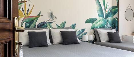 Habitación de hotel con dos camas, un mural de plantas tropicales y un pájaro, y una puerta de madera.