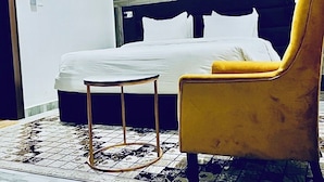 A bedroom with a bed, a yellow armchair, a small round table, and a floral artwork on the wall. - Grand Palmac Hotels (Abuja)