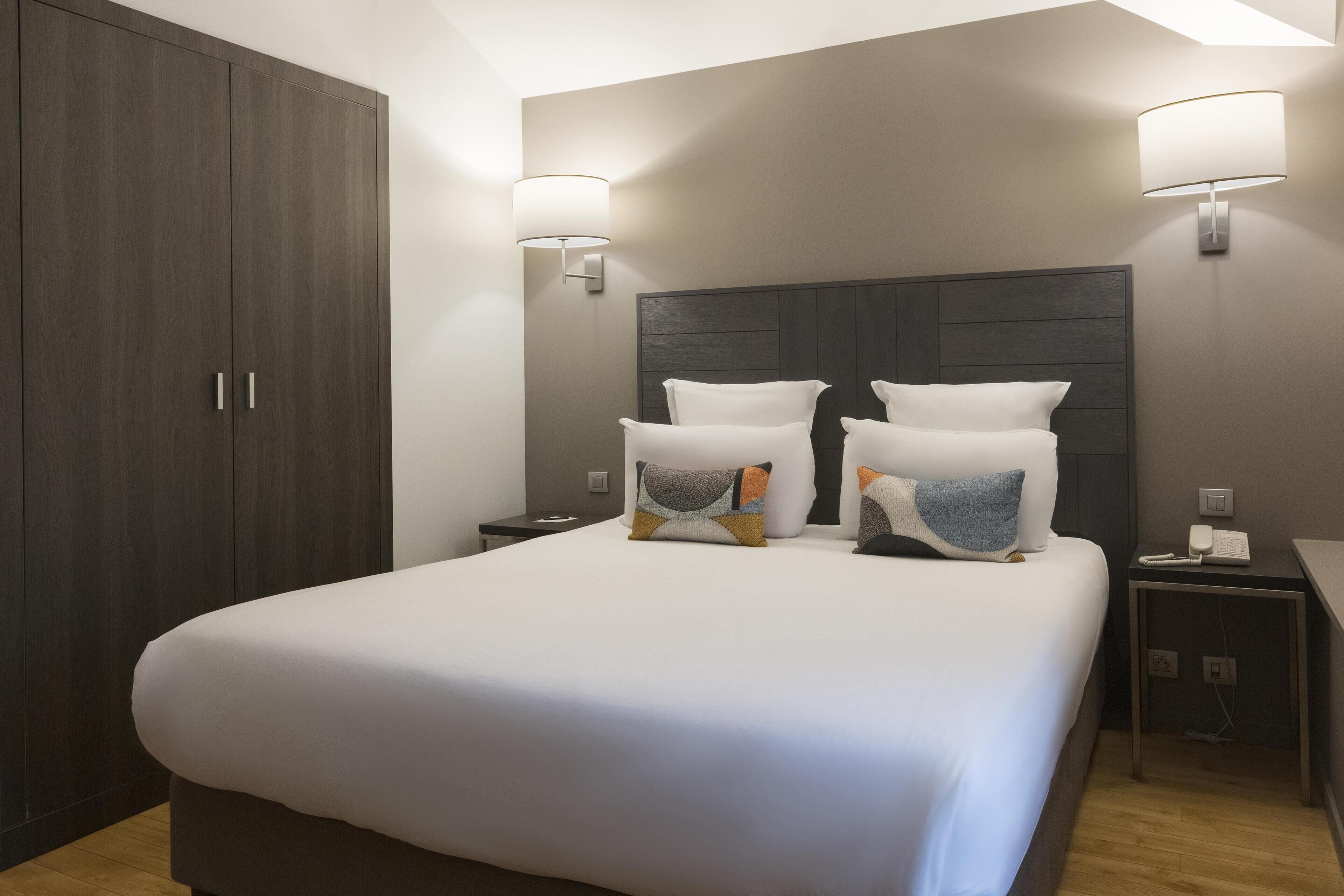 A hotel room with a large bed, two bedside tables, a wardrobe, and wall-mounted lamps.
