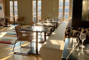 Un espace repas comprenant des tables, des chaises et un canapé, avec de grandes fenêtres donnant sur les bâtiments environnants.