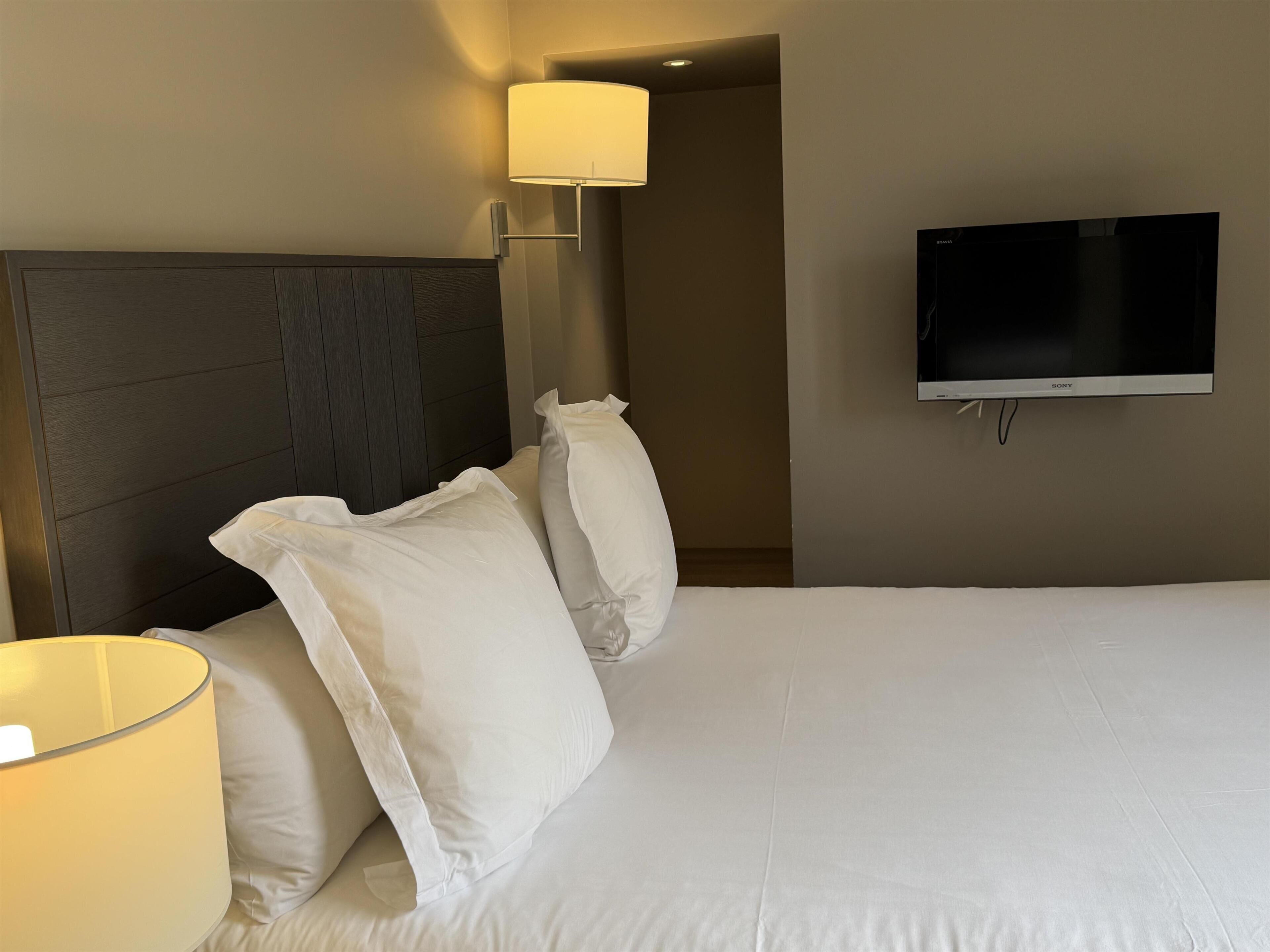A hotel room with a bed, white pillows, a headboard, a wall-mounted TV, and a lamp.