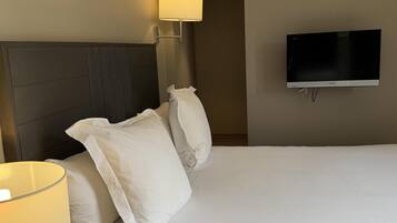 A hotel room with a bed, white pillows, a headboard, a wall-mounted TV, and a lamp.
