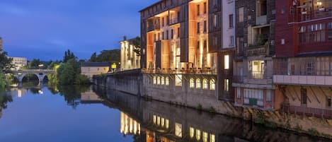 Un edificio junto al río con varios balcones, un puente al fondo y un reflejo tranquilo en el agua.
