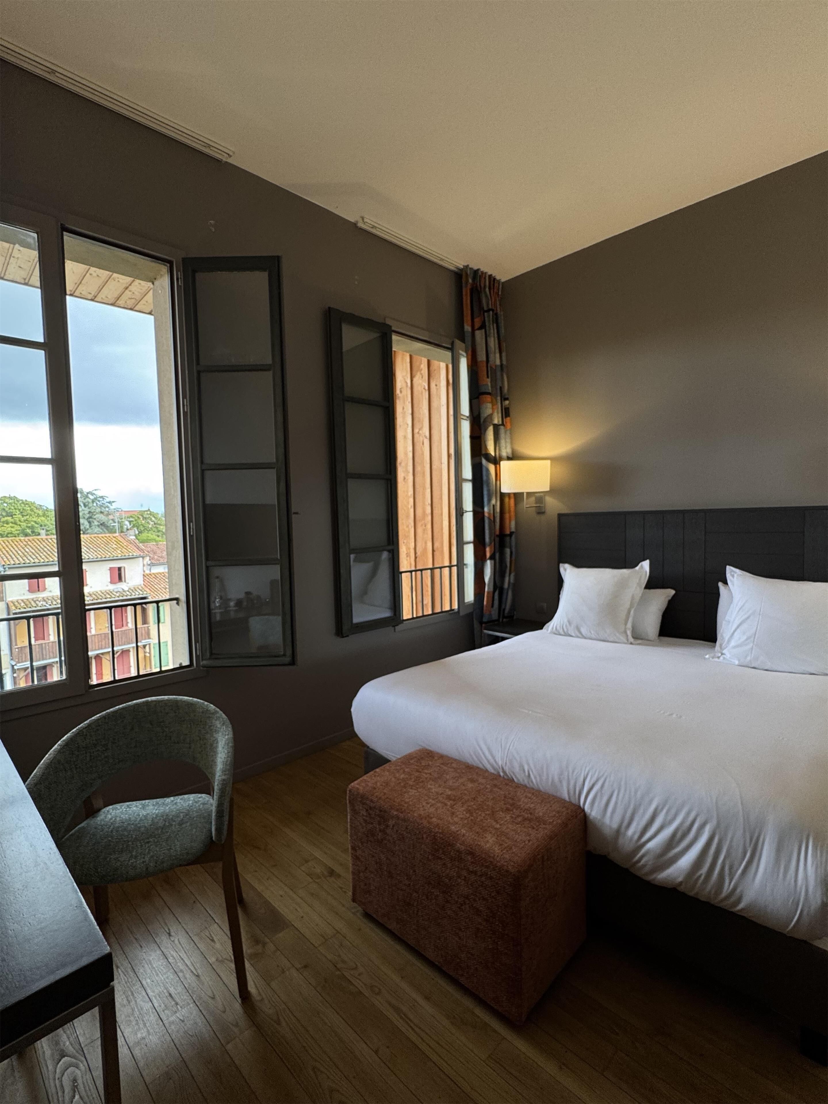 Een hotelkamer met een groot bed, een bureau, een stoel, een raam met uitzicht op gebouwen en een wandlamp.