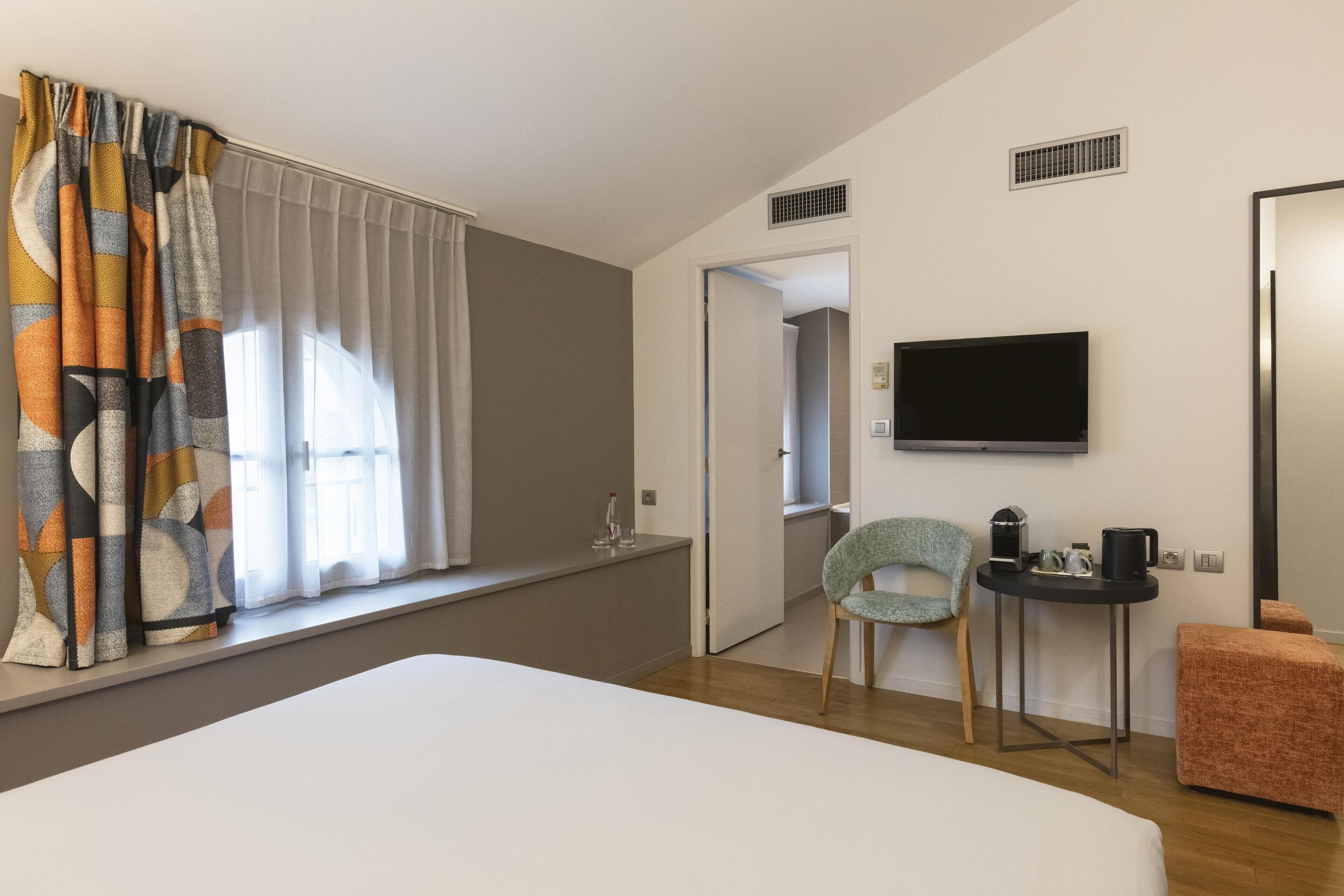 A hotel room with a bed, a television, a small table with a coffee maker, a chair, and a window with curtains.