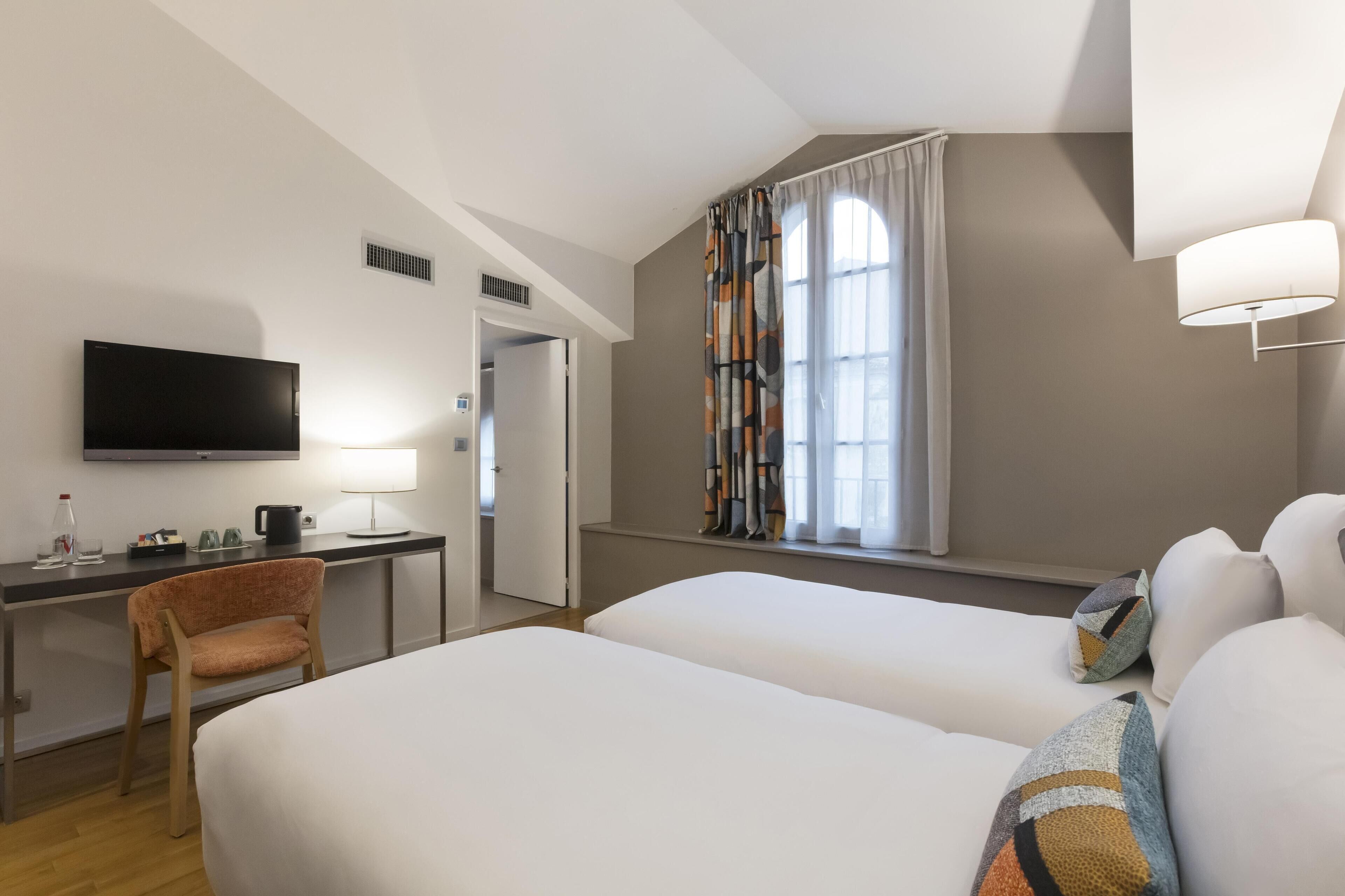 Una habitación de hotel con cama, escritorio con televisor, silla y ventana con cortinas.
