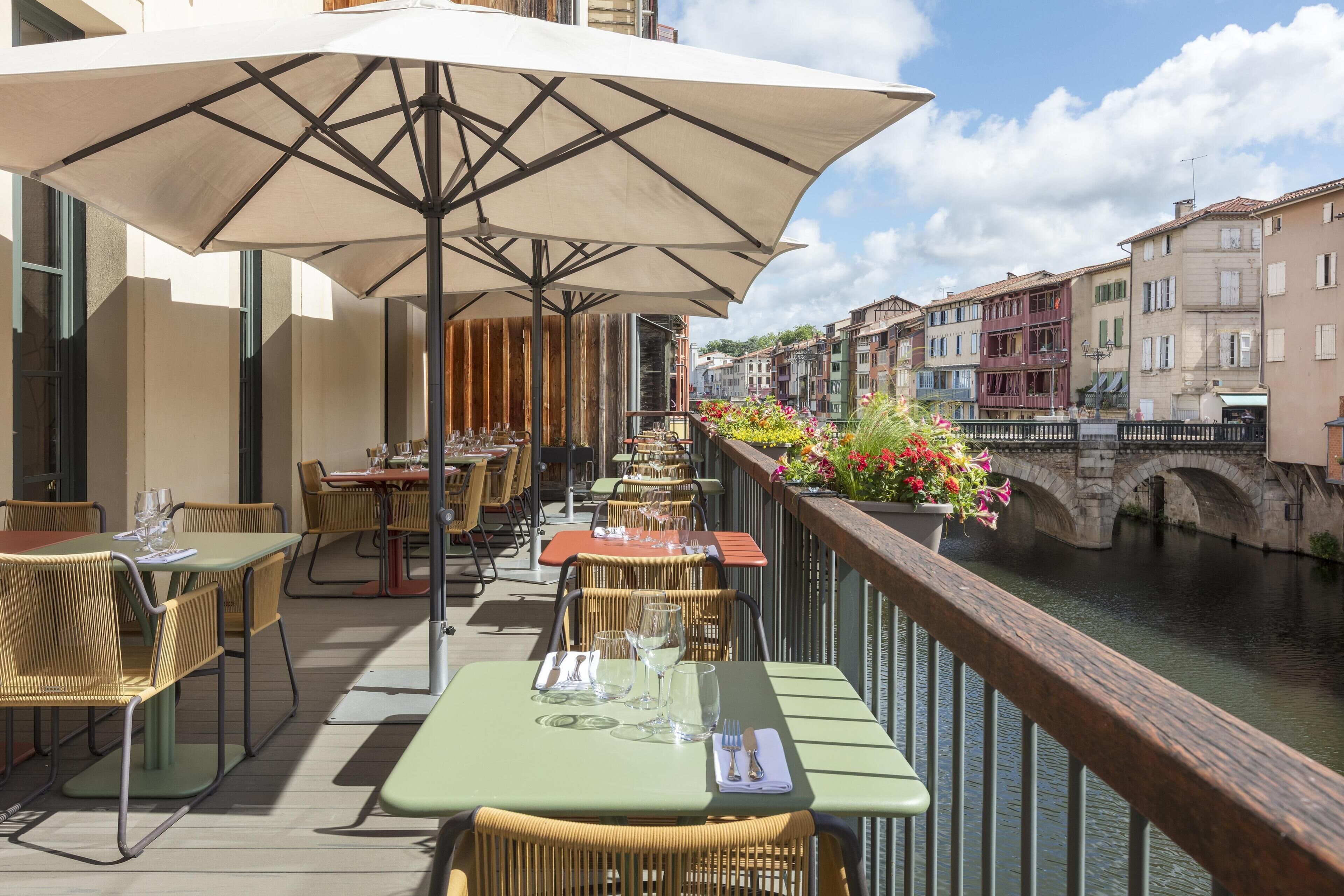 An outdoor dining area with tables and chairs, a railing overlooking a canal, and buildings in the background.