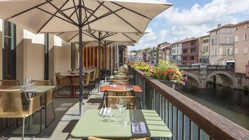 An outdoor dining area with tables and chairs, a railing overlooking a canal, and buildings in the background.