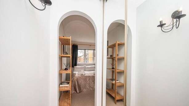 Un couloir étroit mène à une chambre située au bout. La chambre est équipée d’un lit recouvert d’une couvre-lit à motifs, d’une table de chevet en bois et d’une étagère ornée d’objets décoratifs. Le couloir a des murs blancs et un luminaire noir fixé au mur.