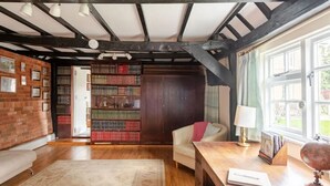 A room with exposed wooden beams, a large bookshelf, a leather sofa, a wooden desk, and a window with a view of greenery. - Cozy Cottage With Scenic Walks (Great Tey)